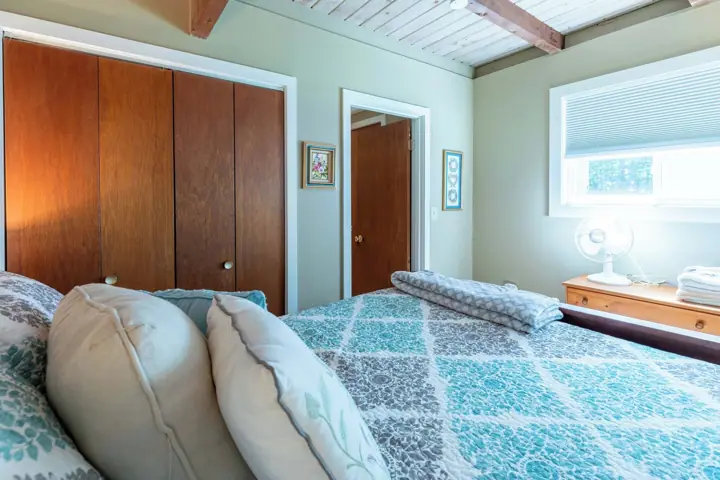 A bedroom with a bed, wooden wardrobe, a bedside table with a fan, and a window with blinds