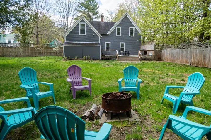 A backyard with a house, a fire pit, and four Adirondack chairs