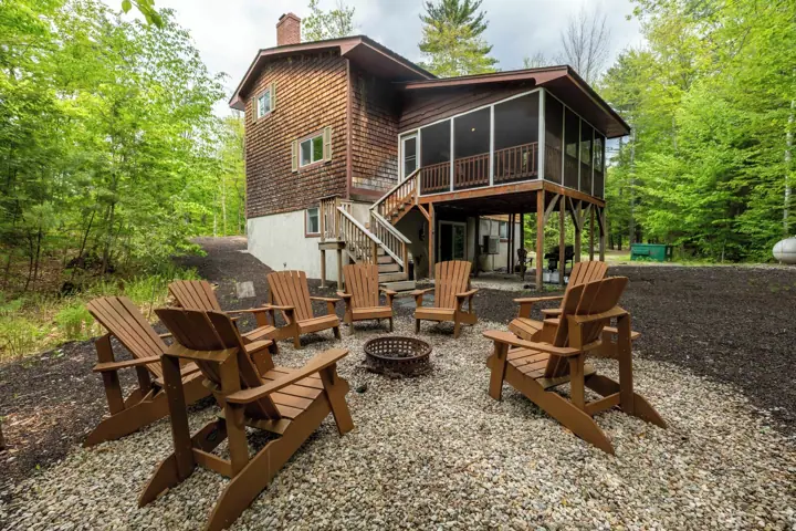 A cabin with a deck, surrounded by Adirondack chairs, a fire pit, and a forested backdrop