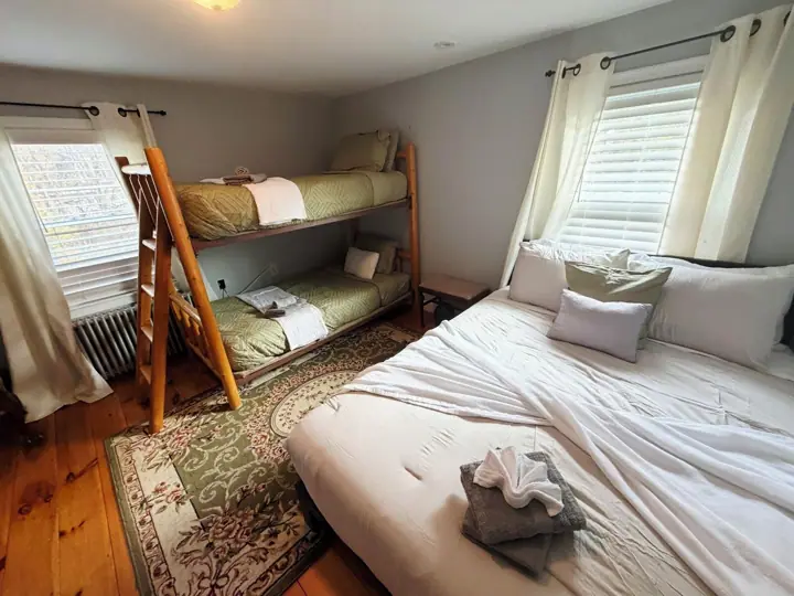 A dormitory room with bunk beds, a wooden ladder, a patterned rug, and a window with curtains