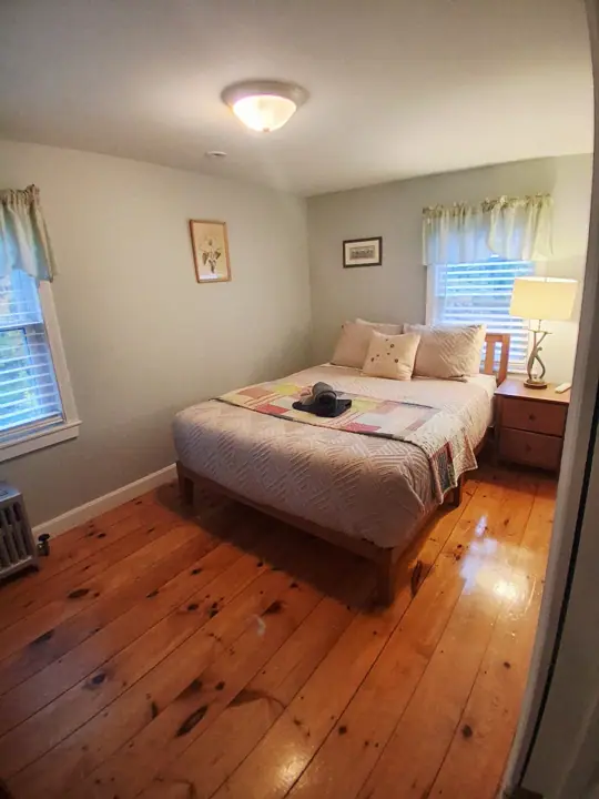 A bedroom with a wooden floor, a bed with a quilt, a nightstand with a lamp, and a window with blinds