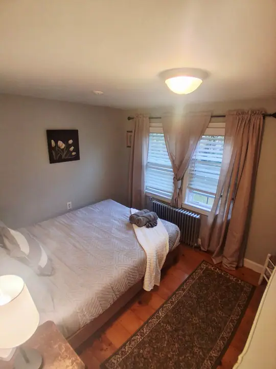A bedroom with a bed, a radiator, a window with curtains, a wall-mounted picture, and a floor lamp