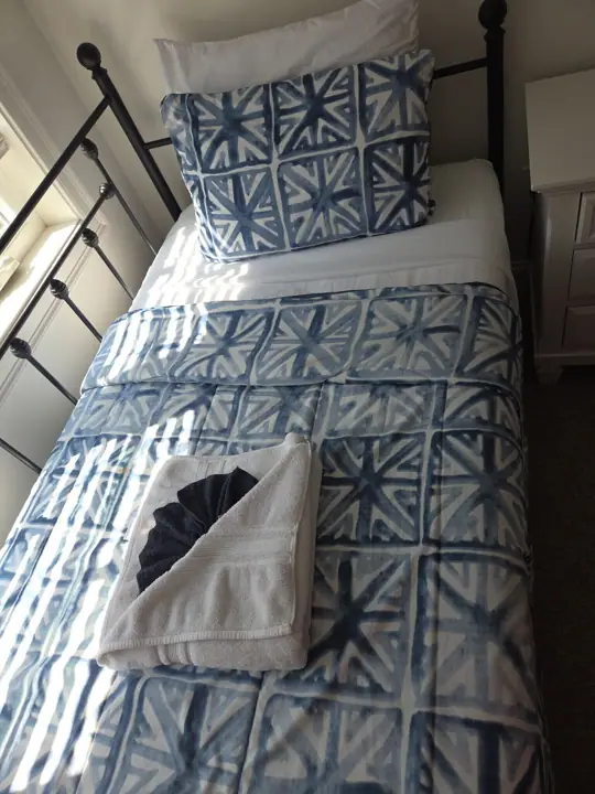 A bed with a patterned bedspread and pillows, a white nightstand, and a window with blinds