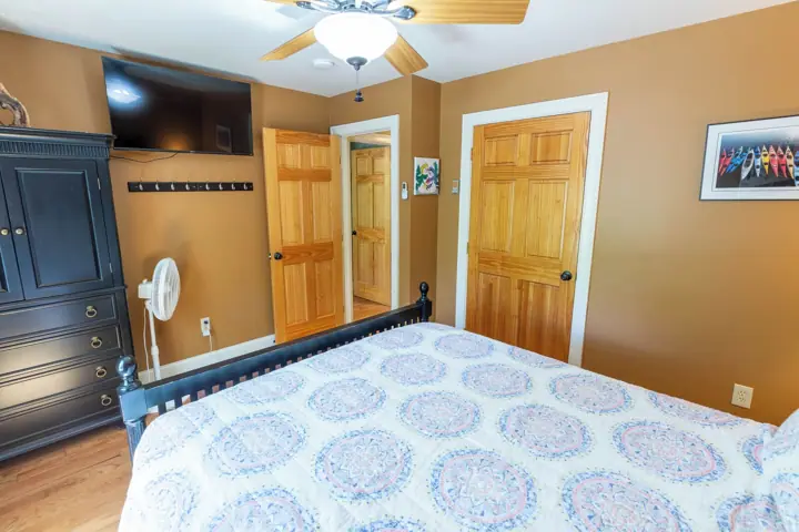 A bedroom with a bed, wooden wardrobe, a ceiling fan, a wall-mounted TV, and a door with a mirror
