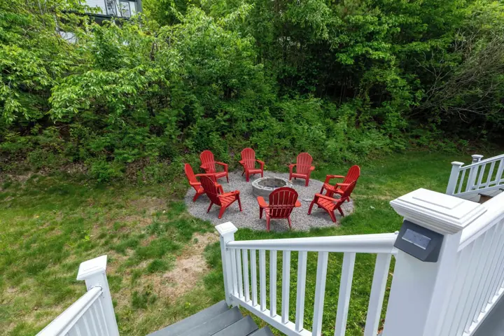 A deck with a fire pit surrounded by red chairs, overlooking a grassy area with dense trees